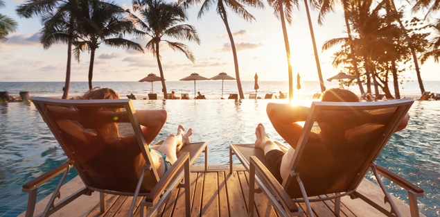 image captured behind two people lounging in reclining chairs on a wooden dock overlooking an ocean and palm trees in the distance while the sun sets