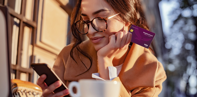 adult female sitting outside in cooler weather looking at her phone while holding a neighborhood cu cash rewards credit card in her other hand