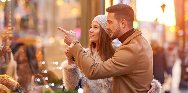 a young couple outdoors wearing late fall clothing to keep warm as they look and point at a window display during the winter holiday season